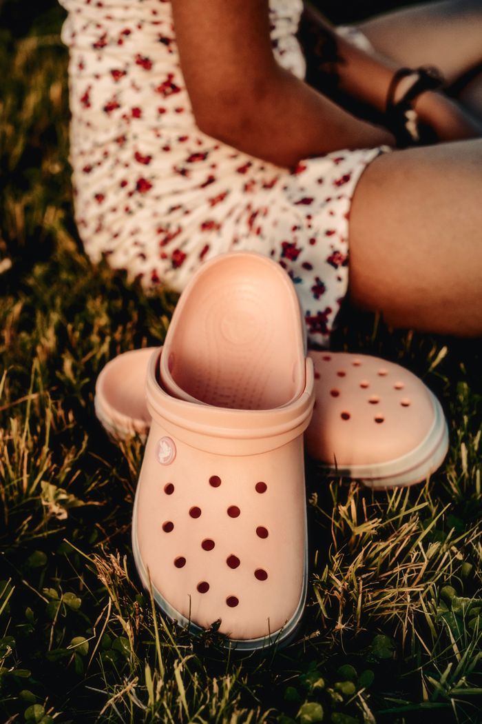 A close-up of pink Crocs on grass beside a woman in a floral dress sitting outdoors.