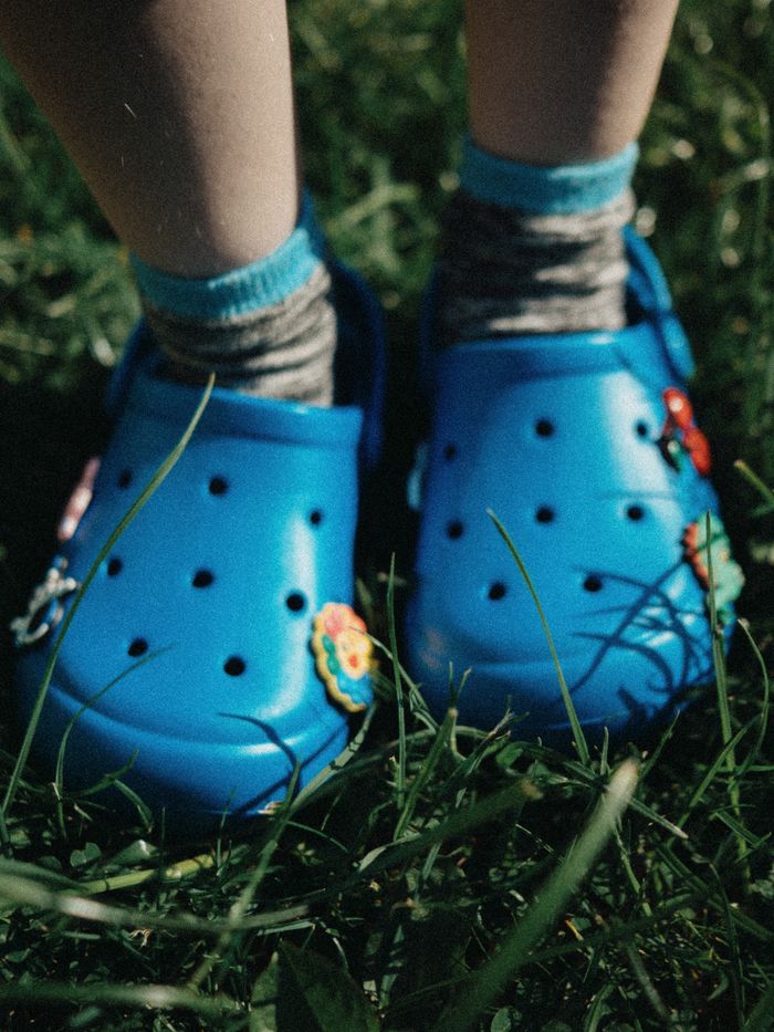 A child's feet in blue Crocs standing on lush green grass during summertime.