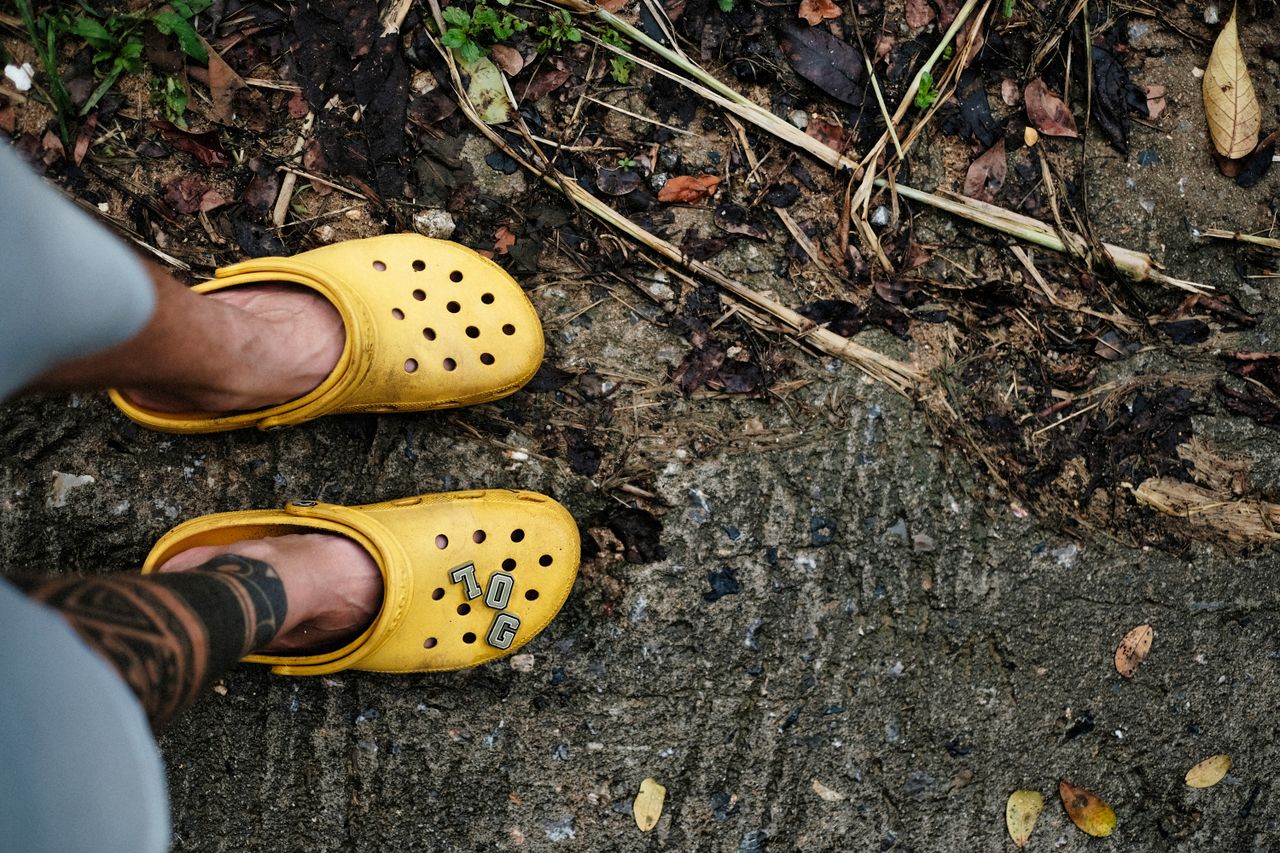 Man in yellow Crocs with tattoo on foot standing on earthy path with dried leaves.