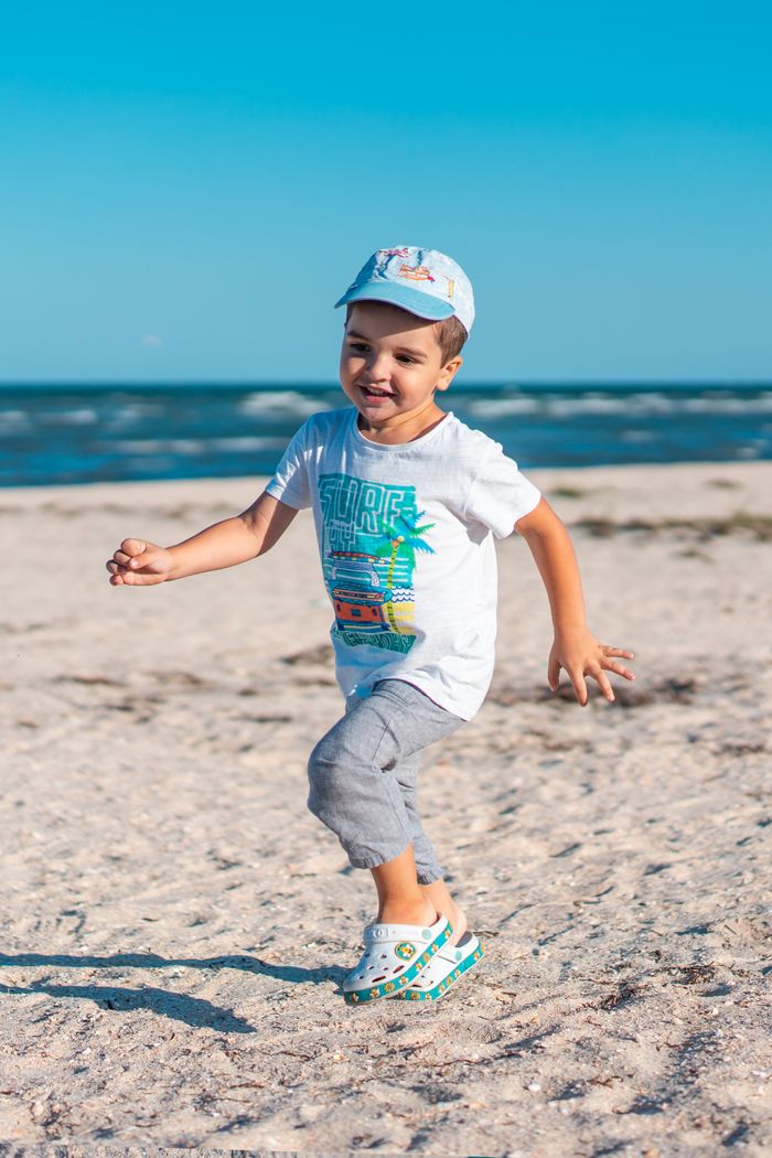 Smiling boy wearing a cap runs on a sandy beach against a clear blue sky.