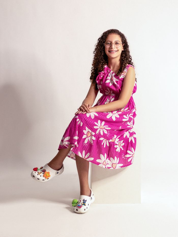 Smiling young girl wearing a pink floral dress with accessorized casual footwear.