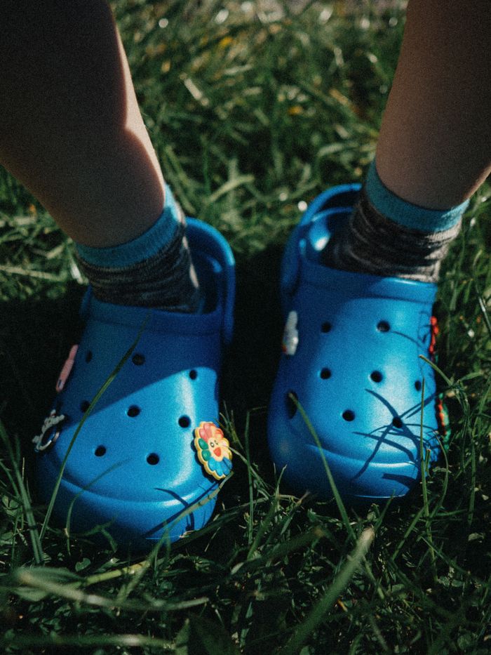 Close-up of a child's feet in blue Crocs on a sunny grassy field.